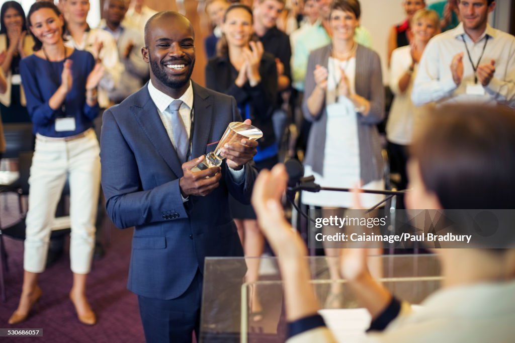 Porträt eines jungen Mannes, der eine Trophäe hält, im Konferenzraum steht und zum applaudierenden Publikum lächelt