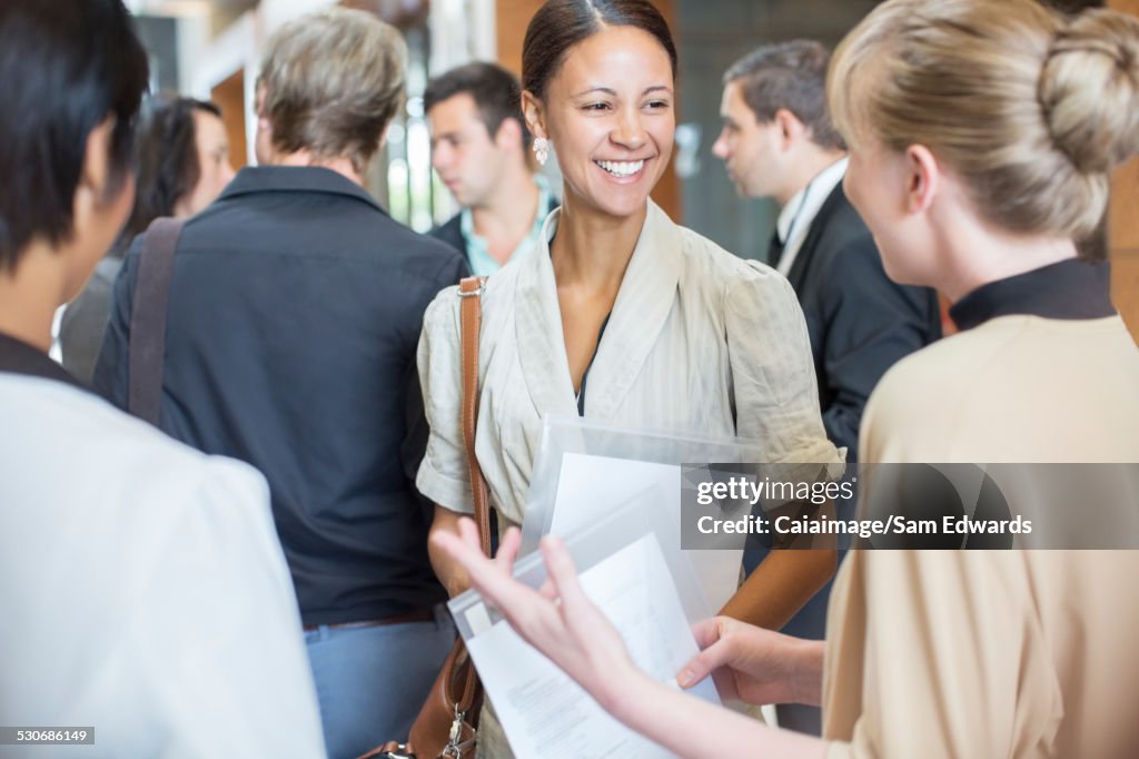 Portrait of two smiling women holding files and talking, standing in crowded lobby