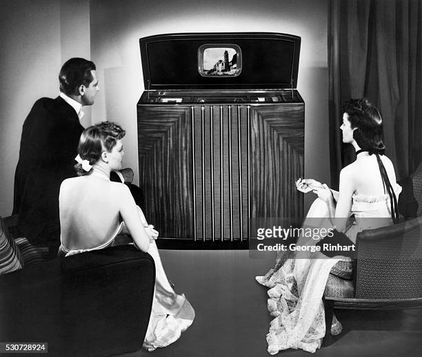 Publicity handout showing formally dressed trio watching large console television. Undated photograph/illustration.