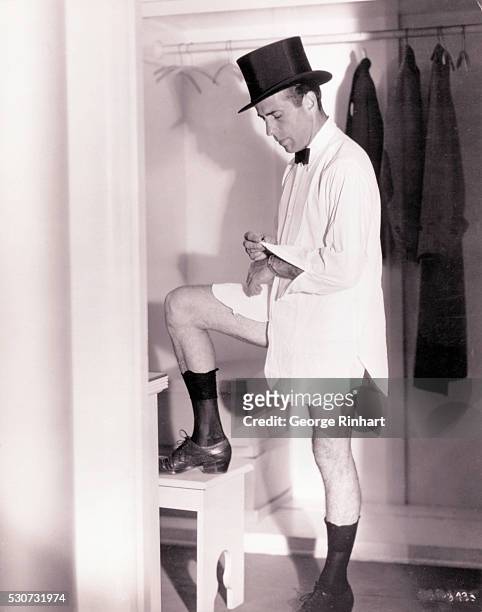 Humphrey Bogart posed for this ludicrous publicity shot in the very young days of his film career. He is shown wearing a top hat, and buttoning the...
