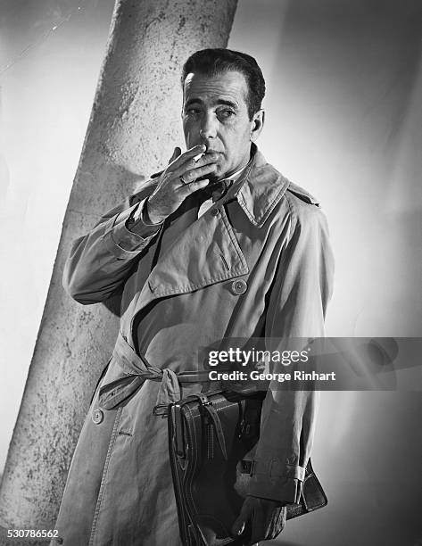 Humphrey Bogart in promotional photo for the film "Sirocco," with Bogey in a classic pose with cigarette in mouth and wearing trench coat. 1951