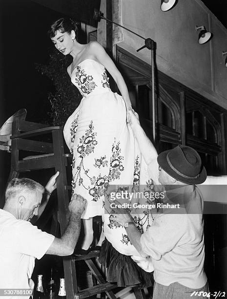 This is a photo of actress Audrey Hepburn standing halfway up a lifeguard's chair as she wears a flower-patterned dress. Two men are helping her.