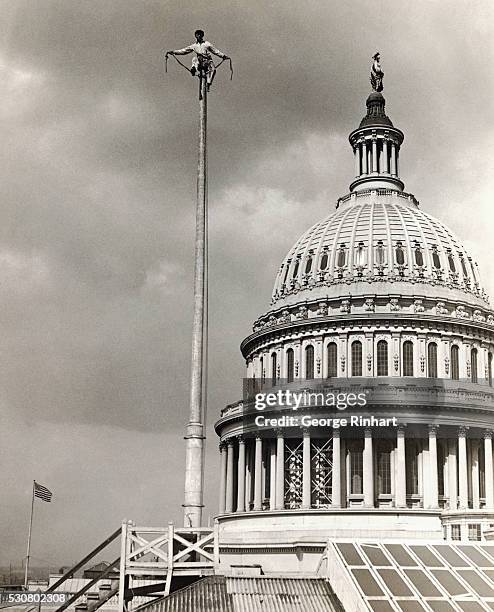 Man sits atop a flagpole that stands alongside the dome of the U. S. Capitol.