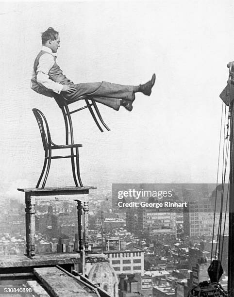 Perched on a girder at the edge of a building under construction, a confident daredevil balances on the back legs of a chair atop another chair set...