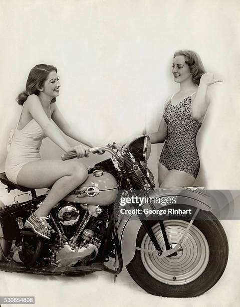 Bathing beauty sits on a Harley-Davidson motorcycle while another woman stands beside. Photograph circa 1940's.