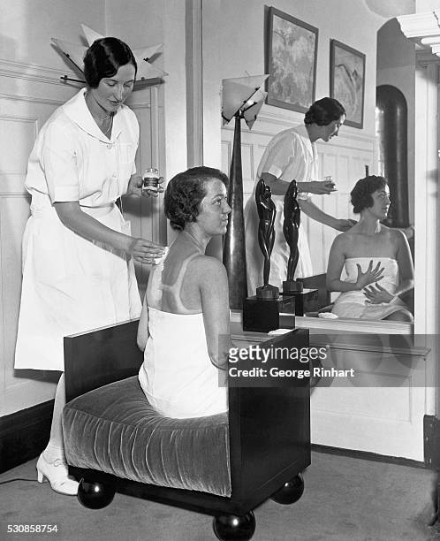 In the fashionable Gold Coast salon of Helena Rubinstein, Carolyn Lang applies lotions to Billie Gloss' suntanned back, bleaching it down so that...