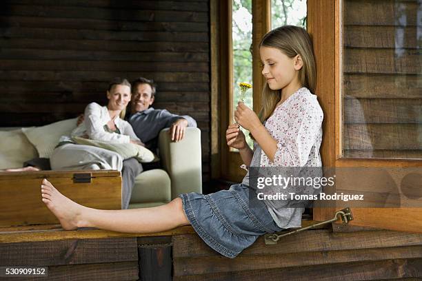 little girl with a flower sitting in a window frame with her parents in the background - wood ledge stock pictures, royalty-free photos & images