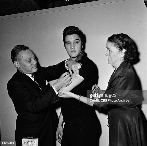 Backstage at the 'The Ed Sullivan Show,' American singer and musician Elvis Presley glances out of the corner of his eye at a smiling nurse while a...