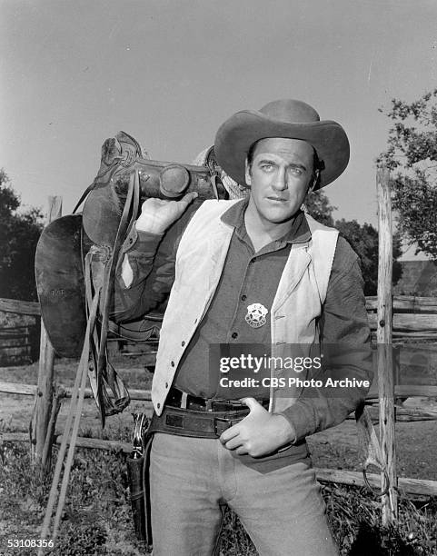 Promotional still of American actor James Arness as he stands near a fence with a saddle swung over one shoulder and his thumb hooked in his belt,...