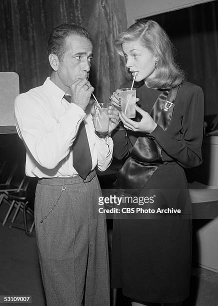 American actors and married couple Humphrey Bogart and Lauren Bacall stare lovingly into each others eyes and sip their drinks through straws while...