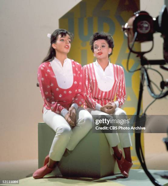 American singer and actress Judy Garland and her daughter, singer and actess Liza Minnelli sit together on a stage cube on top of a raised platform...