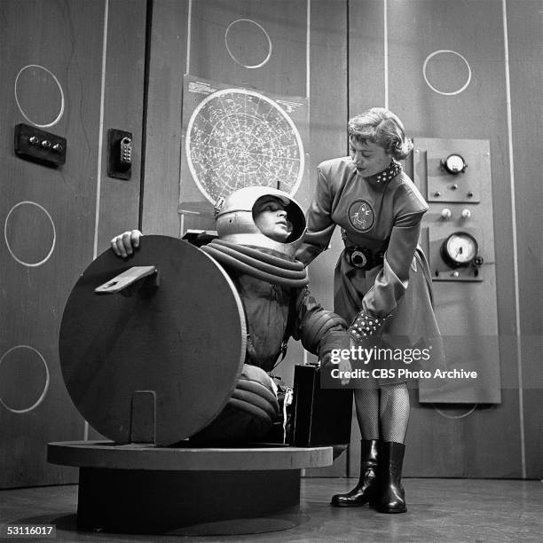 American actress Margaret Garland helps actor Frankie Thomas out of a hatch as he wears a spacesuit and helmet on an episode of the CBS science...