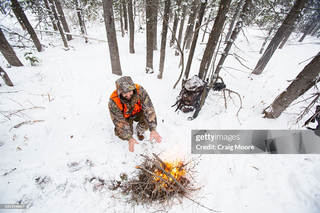 A male hunter stays warm next to a fire in the snow.