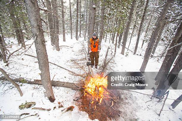 a male hunter stays warm next to a fire in the snow. - spy hunter stock pictures, royalty-free photos & images