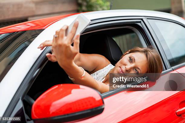 portrait of smiling young woman sitting in her car taking a selfie with her smartphone - stuttgart auto stock pictures, royalty-free photos & images