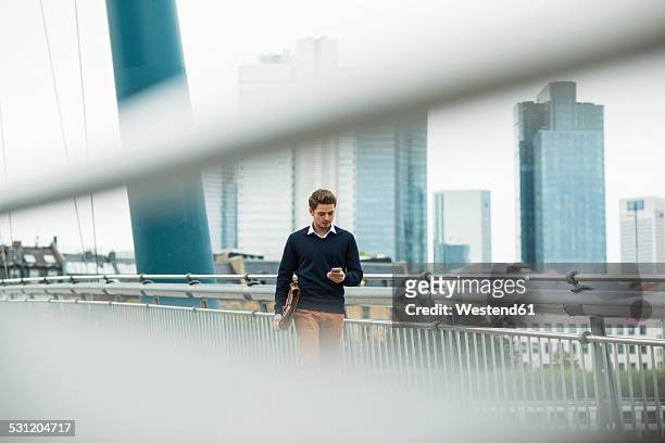 germany, hesse, frankfurt, young businessman walking on a bridge using his smartphone - business man walk stockfoto's en -beelden