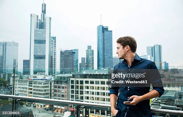 germany, hesse, frankfurt, young man with his smartphone standing in front of the skyline - frankfurt germany skyline stock-fotos und bilder