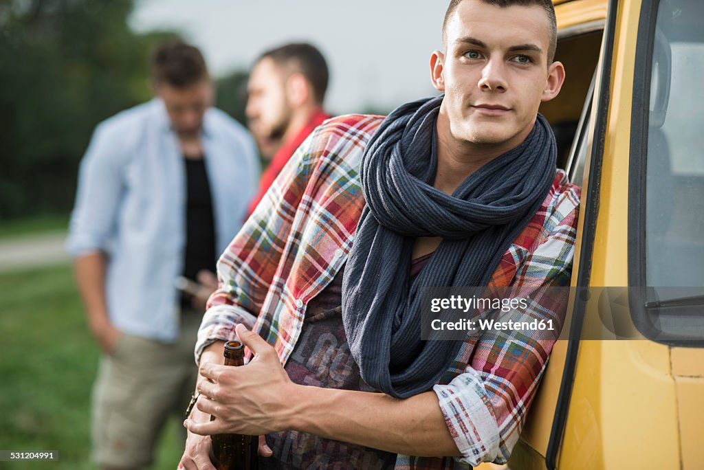 Young man holding bottle of beer leaning against vehicle
