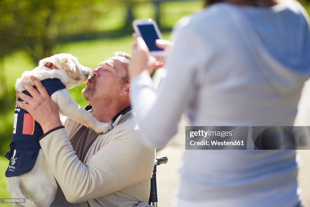 Woman taking picture of man with dog puppy in wheelchair