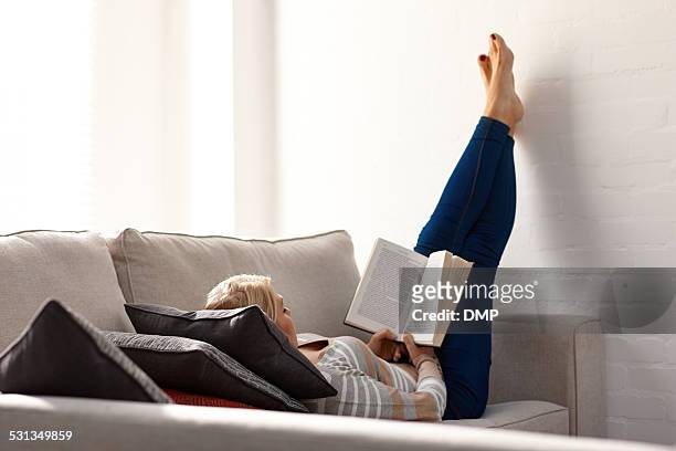 mature woman relaxing on sofa reading a novel - feet up stock pictures, royalty-free photos & images