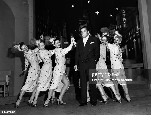 American television personality Ed Sullivan poses, cigarette in hand, as he poses with members of the June Taylor Dancers for a publicity portrait to...