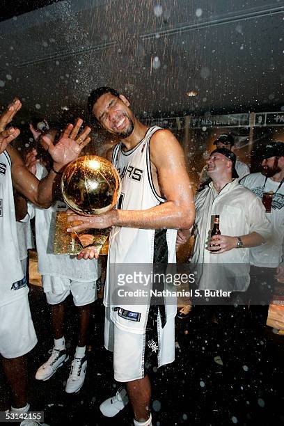 Tim Duncan of the San Antonio Spurs holds the Larry O'Brien NBA Championship trophy as he has champagne sprayed on him by teammates in the locker...