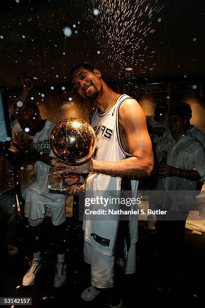 Tim Duncan of the San Antonio Spurs holds the Larry O'Brien NBA Championship trophy as he has champagne sprayed on him by teammates in the locker...