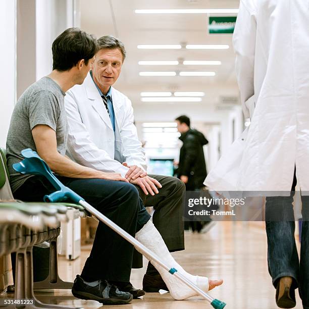 man with cruches and cast on broken leg consulting doctor - injured man in hospital bed stockfoto's en -beelden