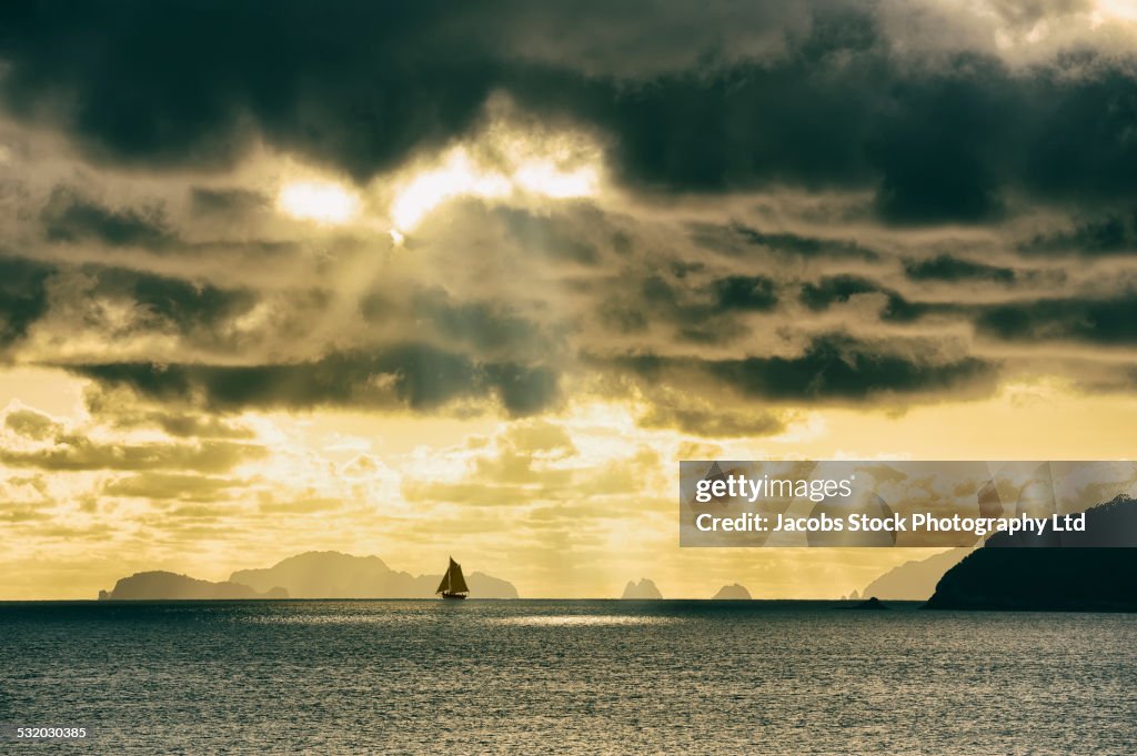 Sun streaming through dark storm clouds over sailboat on ocean