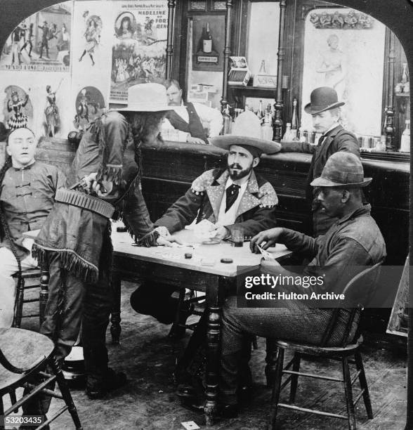 Man in a fringed leather jacket and cowboy hat holds a revolver at his waist as he argues with a seated fellow gambler in a staged photograph...