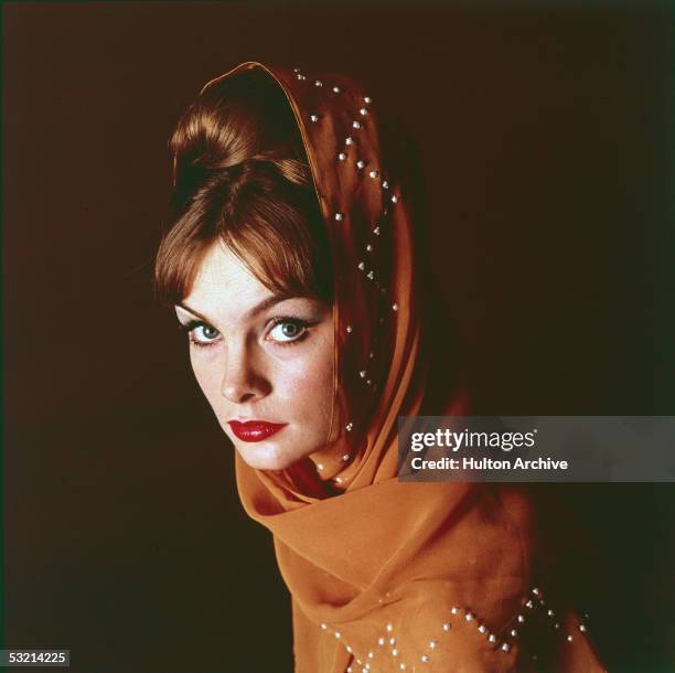 English fashion model Jean Shrimpton poses, looking at the camera, with a pumpkin-colored beaded scarf wrapped around her bouffant hairdo, 1960s.