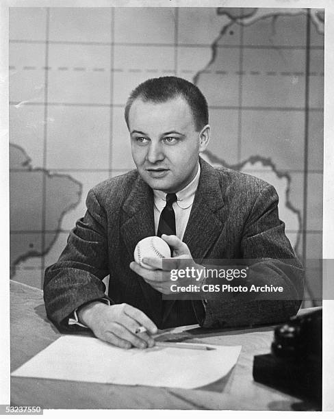 American sports journalist and television personality Jim McKay sits at a desk and holds up a baseball in his left hand while he smokes a cigarette ,...