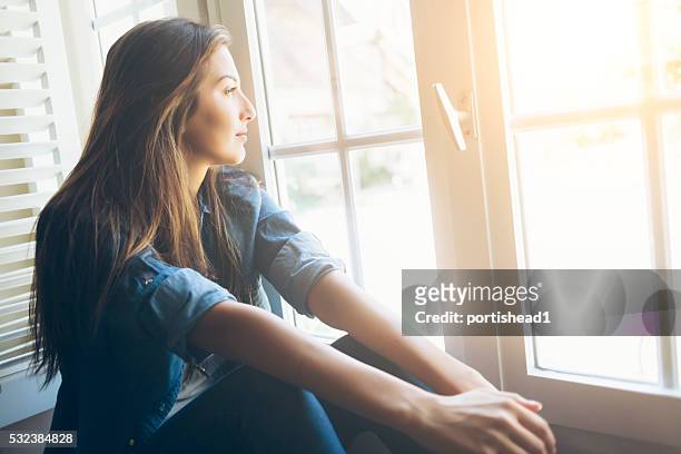 young woman sitting and looking through window - sun rays through window stock pictures, royalty-free photos & images