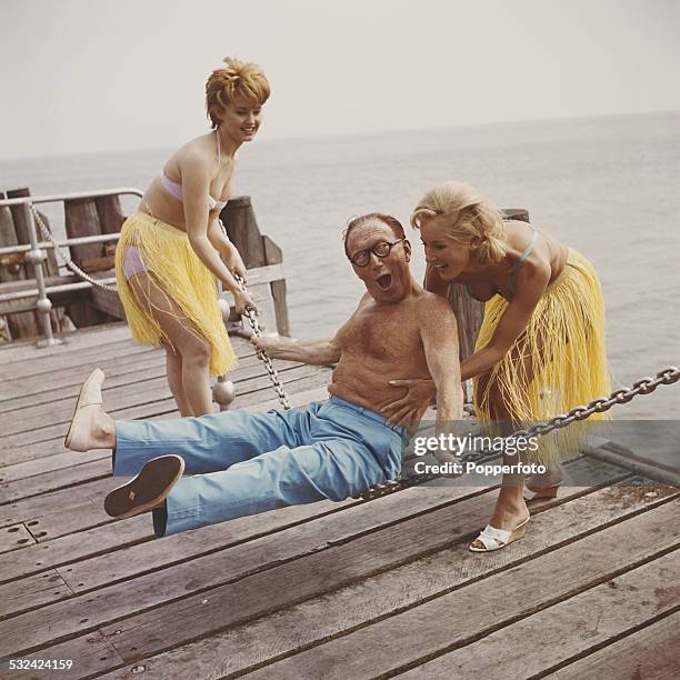 English actor and comedian, Arthur Askey pictured bare chested with two young girls wearing bikinis and grass hula skirts, swinging him on a metal...