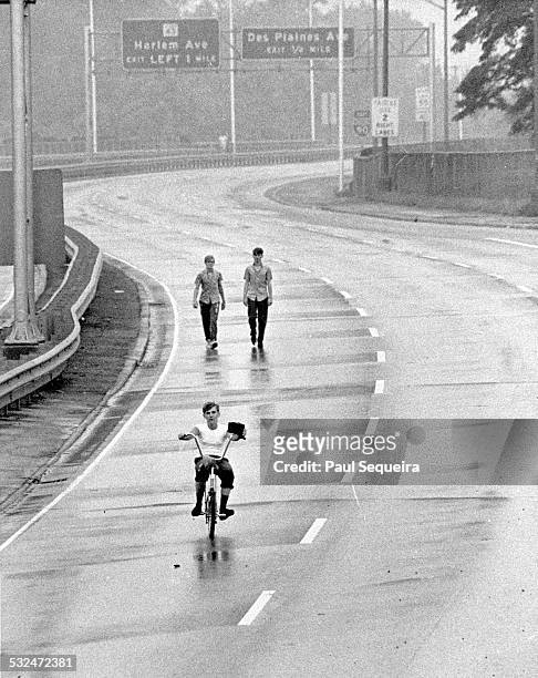 Boy holds a radio as he rides a bicycle along a lane on the closed Eisenhower Expressway, Chicago, Illinois, 1968. Two others walk behind him. The...