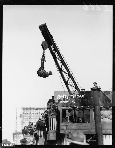 Crowd watches Hungarian-born American magician, escape artist, and psychic debunker Harry Houdini as he wears a straitjacket and hangs from a crane...