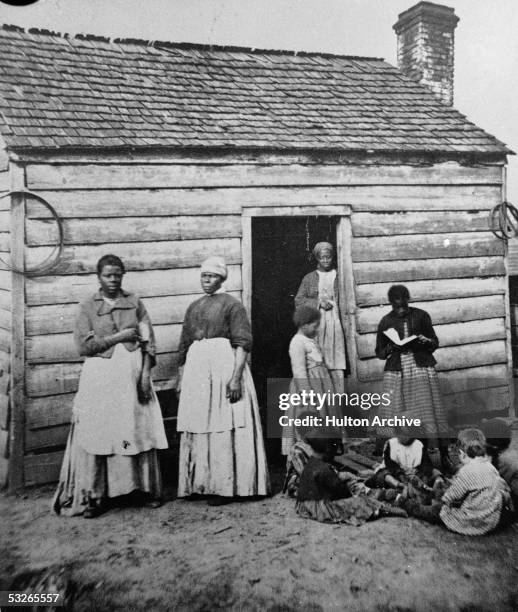 Group of women and children, presumably enslaved, sit and stand around the doorway of a rough wooden cabin, Southern United States, mid 19th Century....