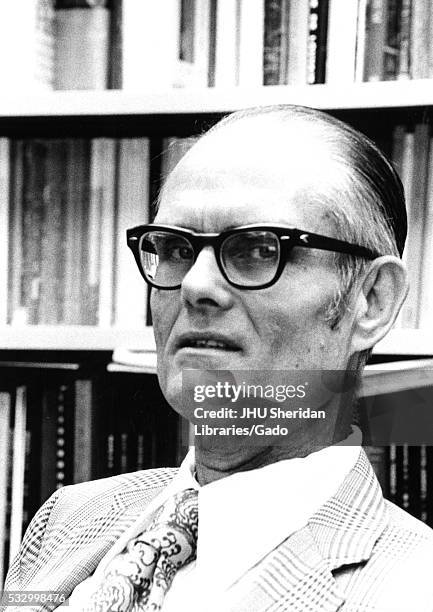 Head-shot of psychologist Julian Stanley, wearing a patterned suit and tie with a white shirt and dark thick glasses, seated in front of a bookshelf...