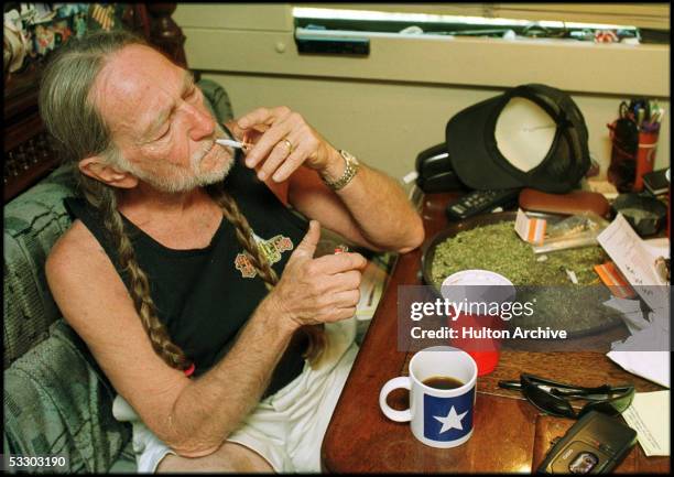 American country singer Willie Nelson takes a drag off a joint while relaxing at his home in Texas, 2000s. A large amount of marijuana is spread out...