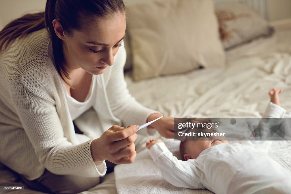 Worried mother measuring baby's temperature in bedroom.