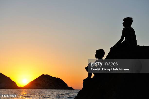 two silhouetted boys sitting on a rock in the sea at sunset - petani stock pictures, royalty-free photos & images