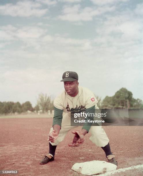 American professional baseball player Jackie Robinson of the Brooklyn Dodgers, dressed in a road uniform, crouches by the base and prepares to catch...