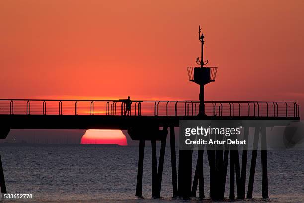the sunrise moment on the mediterranean sea with the silhouette of people walking on a long dock with the sun emerging from the horizon with nice colors and shapes. - badalona stock pictures, royalty-free photos & images