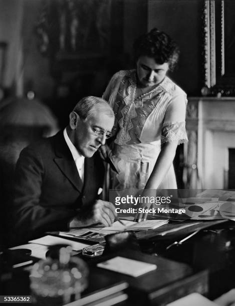 American politician Woodrow Wilson , President of the United States from 1913-1921, goes over papers at his desk as his second wife Edith Bolling...