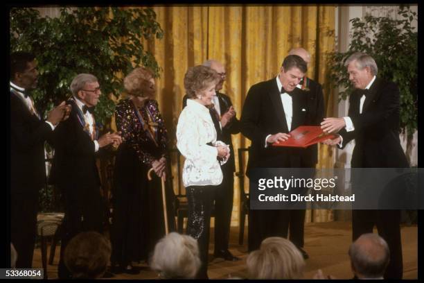 Former President Ronald Reagan receiving award from Ralph Davidson with other Kennedy Center honorees Loy, Burns & Ailey applauding.