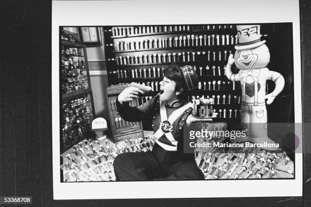 Pezhead collector Sue Sternfeld in self-made Pez suit holding plastic gun to her mouth & tray filled with Pezheads in her Pezhead crowded living room.