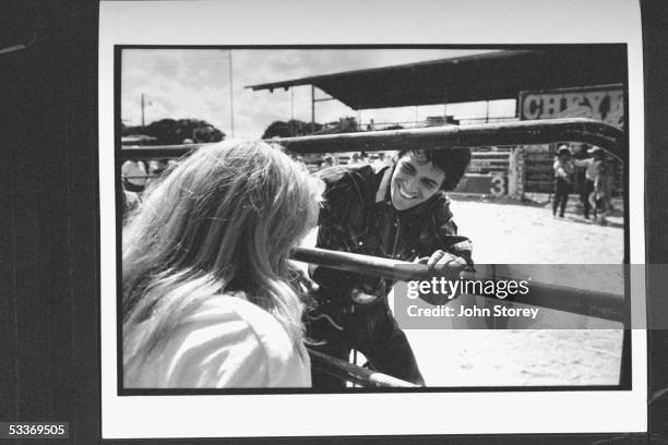 Actor Luke Perry standing by iron railing inside rodeo ring as he chats with female fan who is standing outside ring during a break in the filming of...