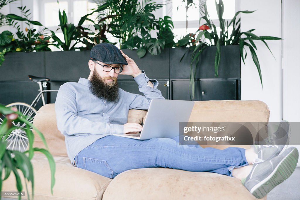 Bärtiger hipster Lehnen Sie sich auf der couch und arbeiten auf laptop-Computer