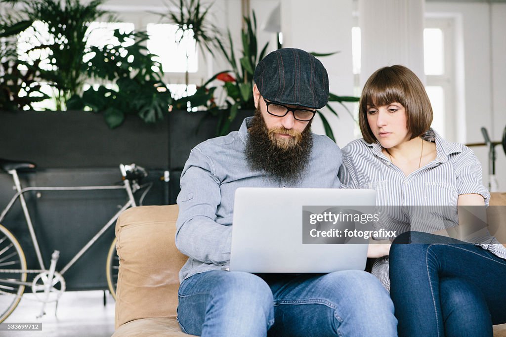 Hipster and girlfriend sit on couch and work at laptop