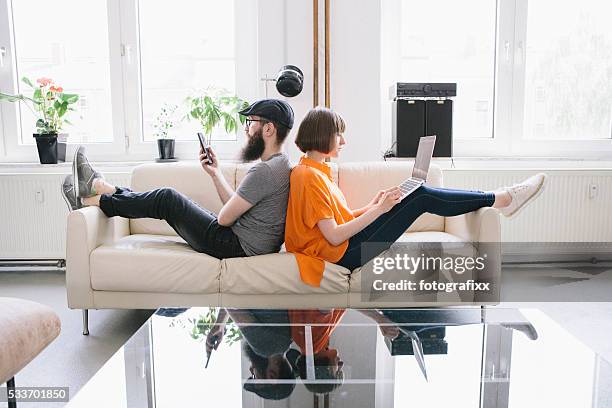 hipster couple sit on couch and look into digital devices - couple sitting stock pictures, royalty-free photos & images